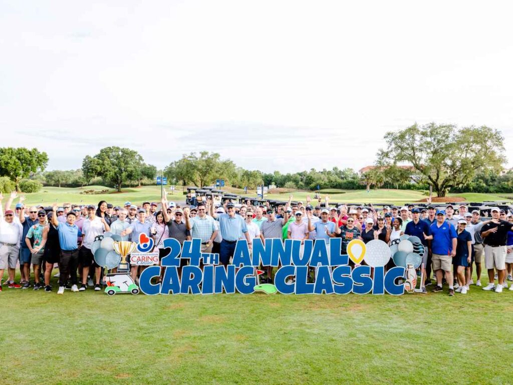 A large group of golfers happily standing behind the Caring Classic golf event sign.